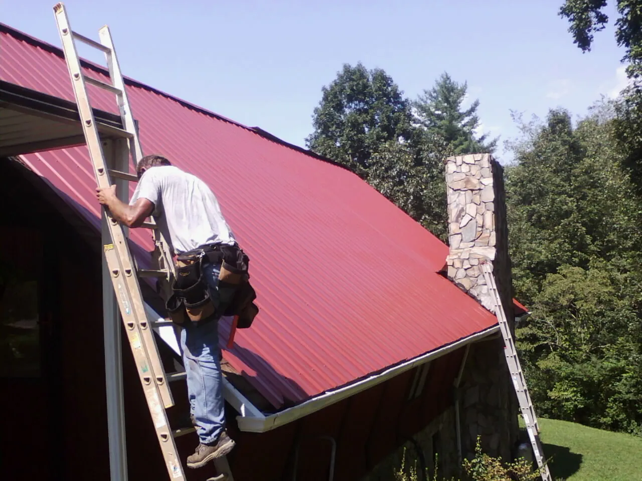 Roofer on a ladder beside a red metal roof and stone chimney — Soffit & Fascia Repair by Roofing Brothers in Hermanville
